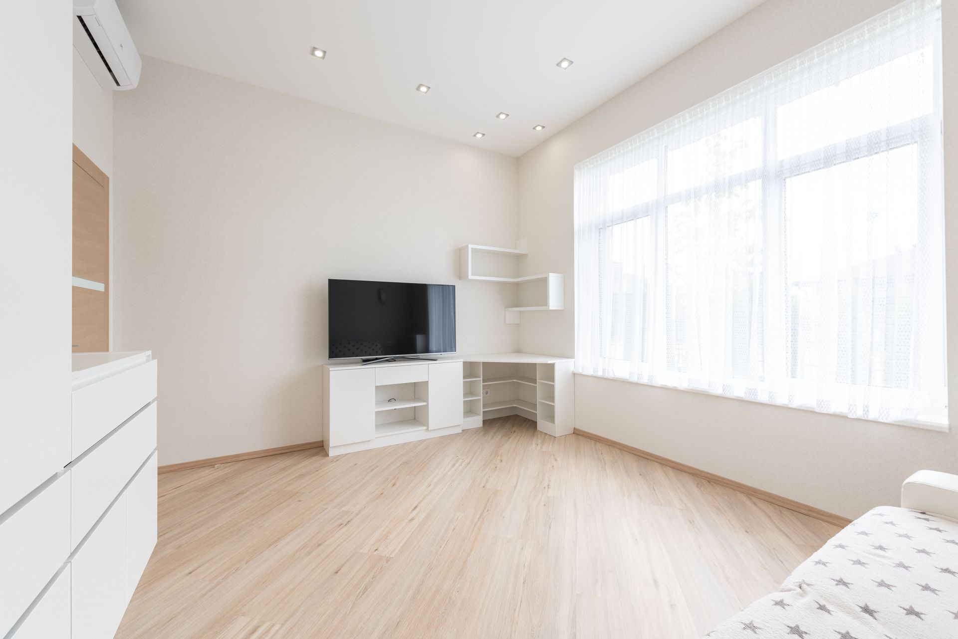 Modern TV set on cupboard placed near empty table in spacious room with big window and white cabinets in apartment